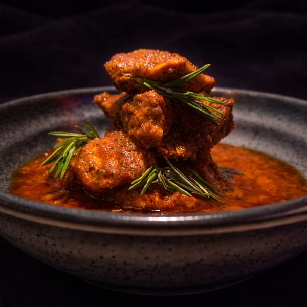 Spicy chicken pieces in a rich red sauce, garnished with fresh rosemary in a dark bowl.
